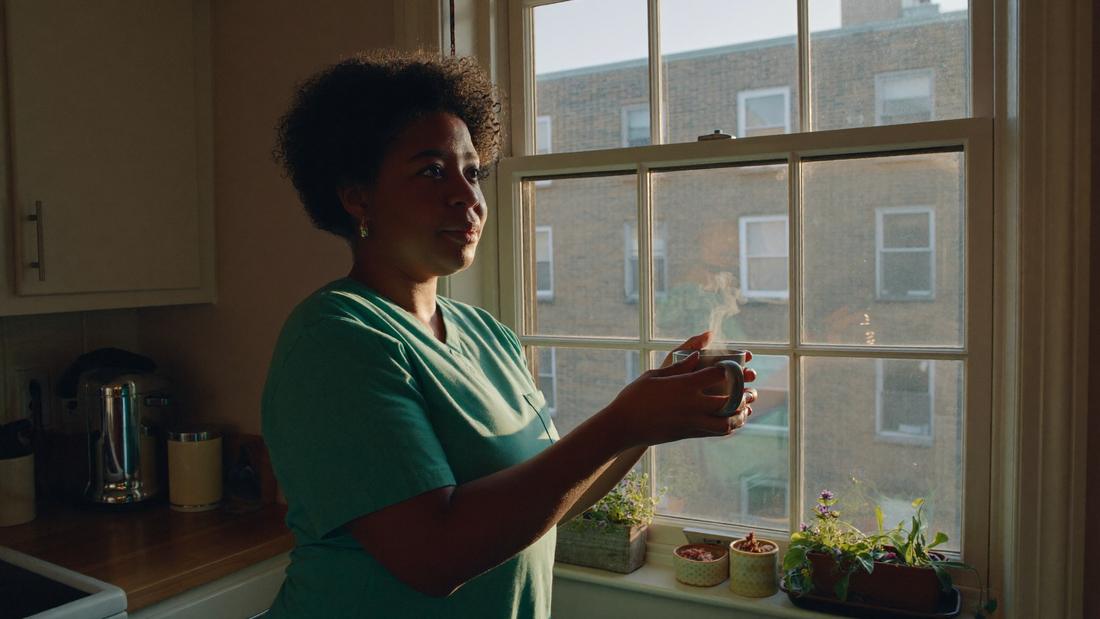 A nurse stands at her own kitchen window with a cup of coffee at dawn, quiet and unhurried.